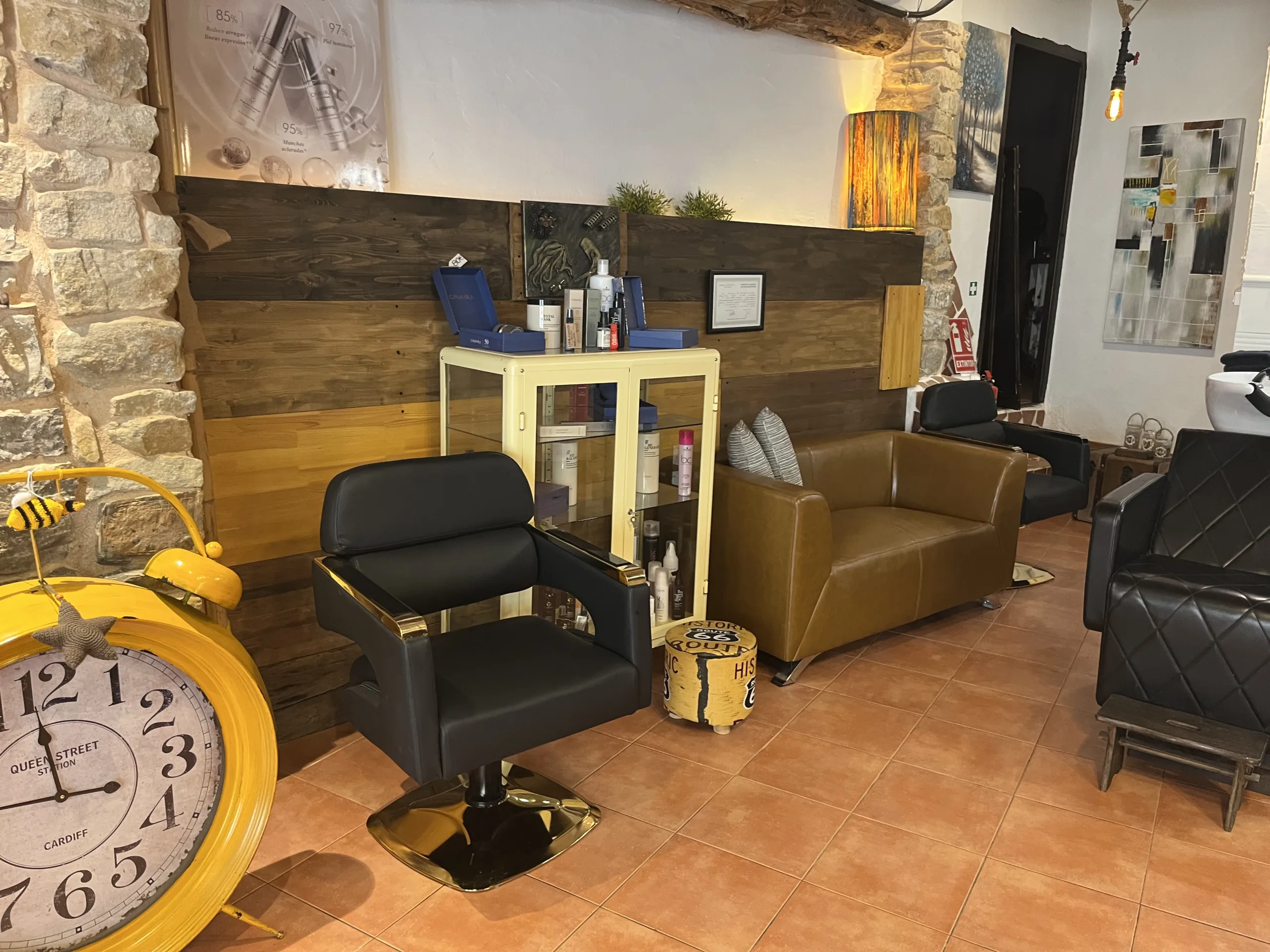 Zona de espera en salón de belleza con sillón marrón, silla de peluquería negra, vitrina de productos y reloj decorativo amarillo.