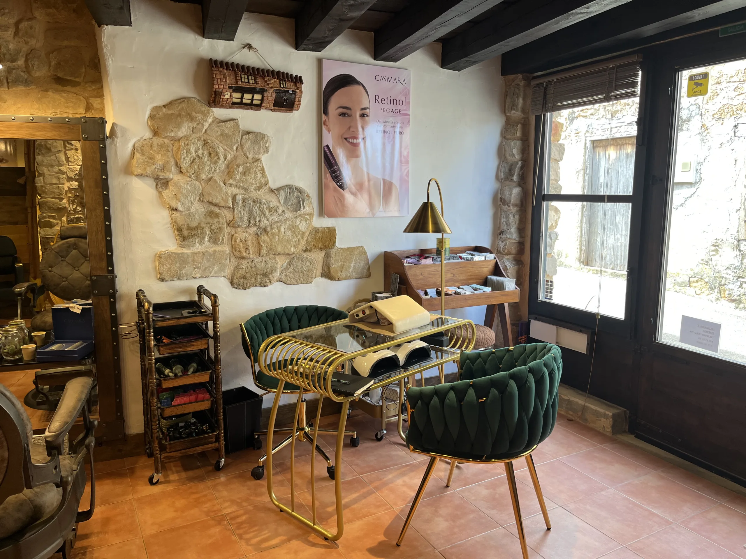 Zona de manicura con mesa dorada, dos sillas verdes aterciopeladas, cartel publicitario y decoración rústica con piedra vista.
