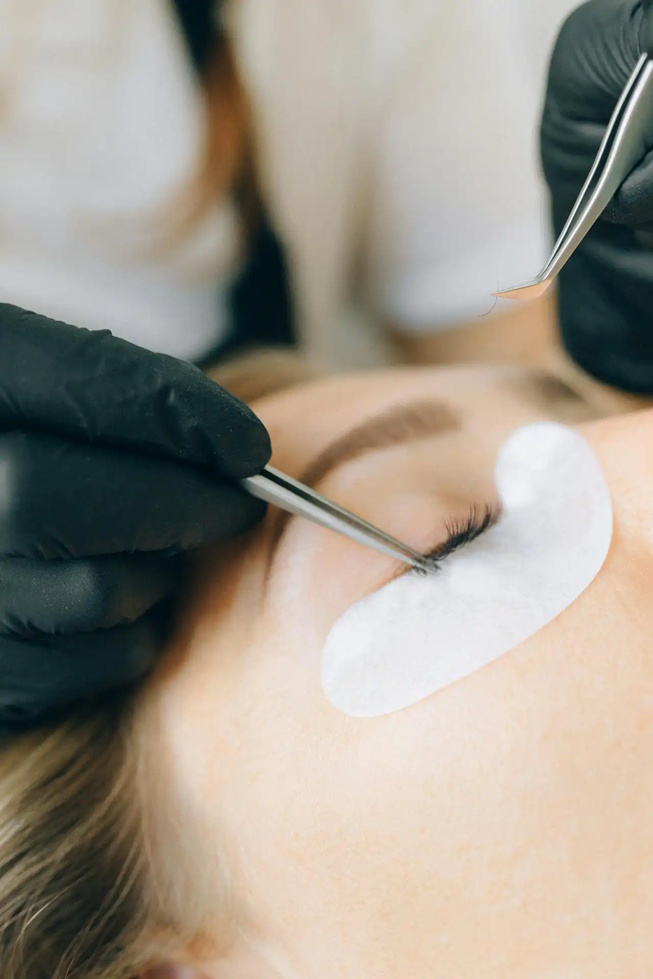 Aplicación de extensiones de pestañas con pinzas, sobre parche ocular blanco.