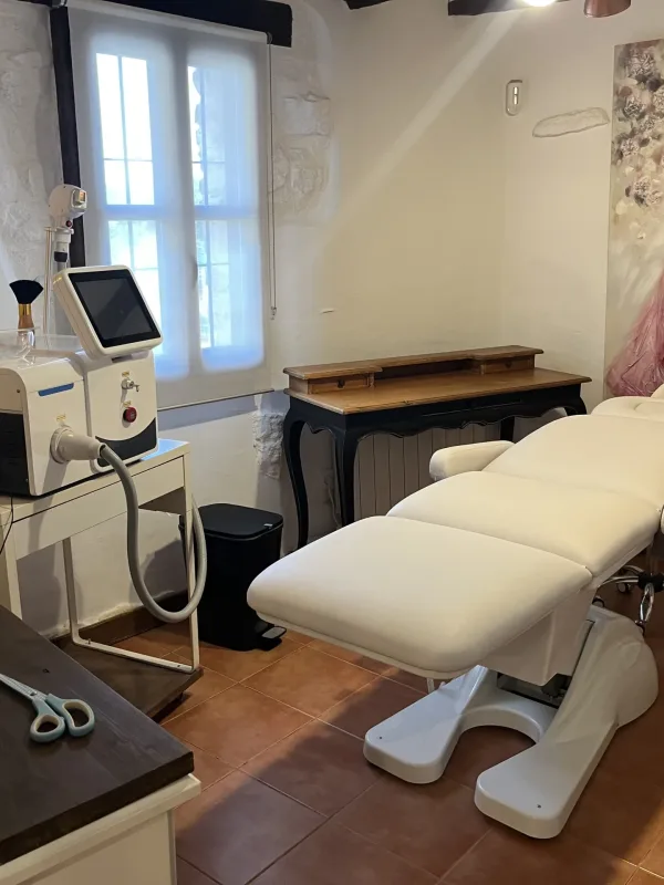 Cabina estética con camilla blanca, mobiliario rústico y decoración con cuadro de vestido rosa.