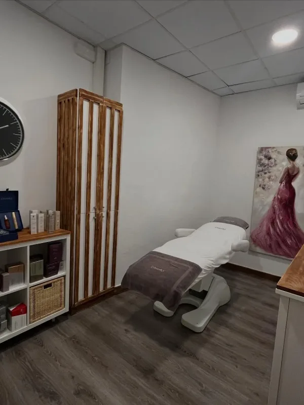 Sala de estética moderna con camilla blanca, estantería de productos y cuadro de vestido rosa.